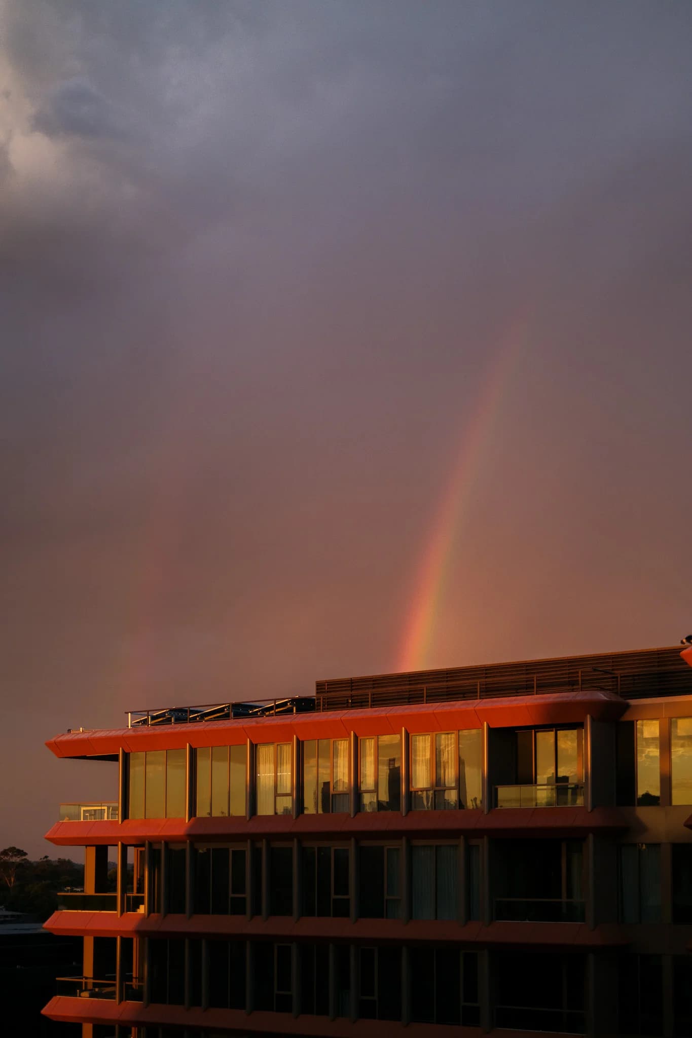 A distinct rainbow appears above a modern building with red-orange features. Golden hour light reflects in the building's windows against a dark sky.