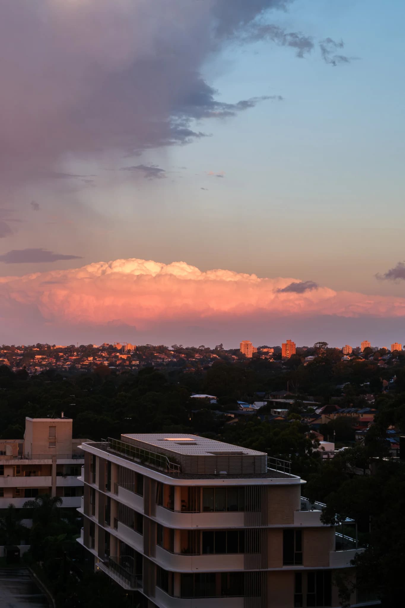 Large, pink-hued clouds float above a city's rooftops illuminated by the setting sun. A modern building with curved facades occupies the foreground.