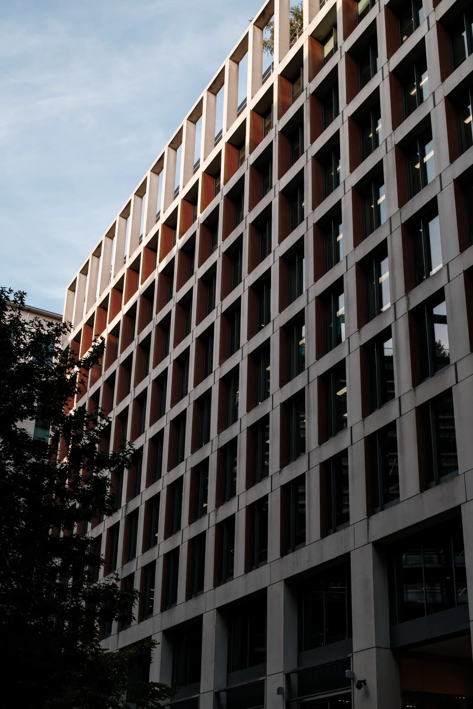 A modern building displays a grid pattern of concrete frames and dark windows under a blue sky. Sunlight illuminates the upper sections, highlighting the repeated structural elements.