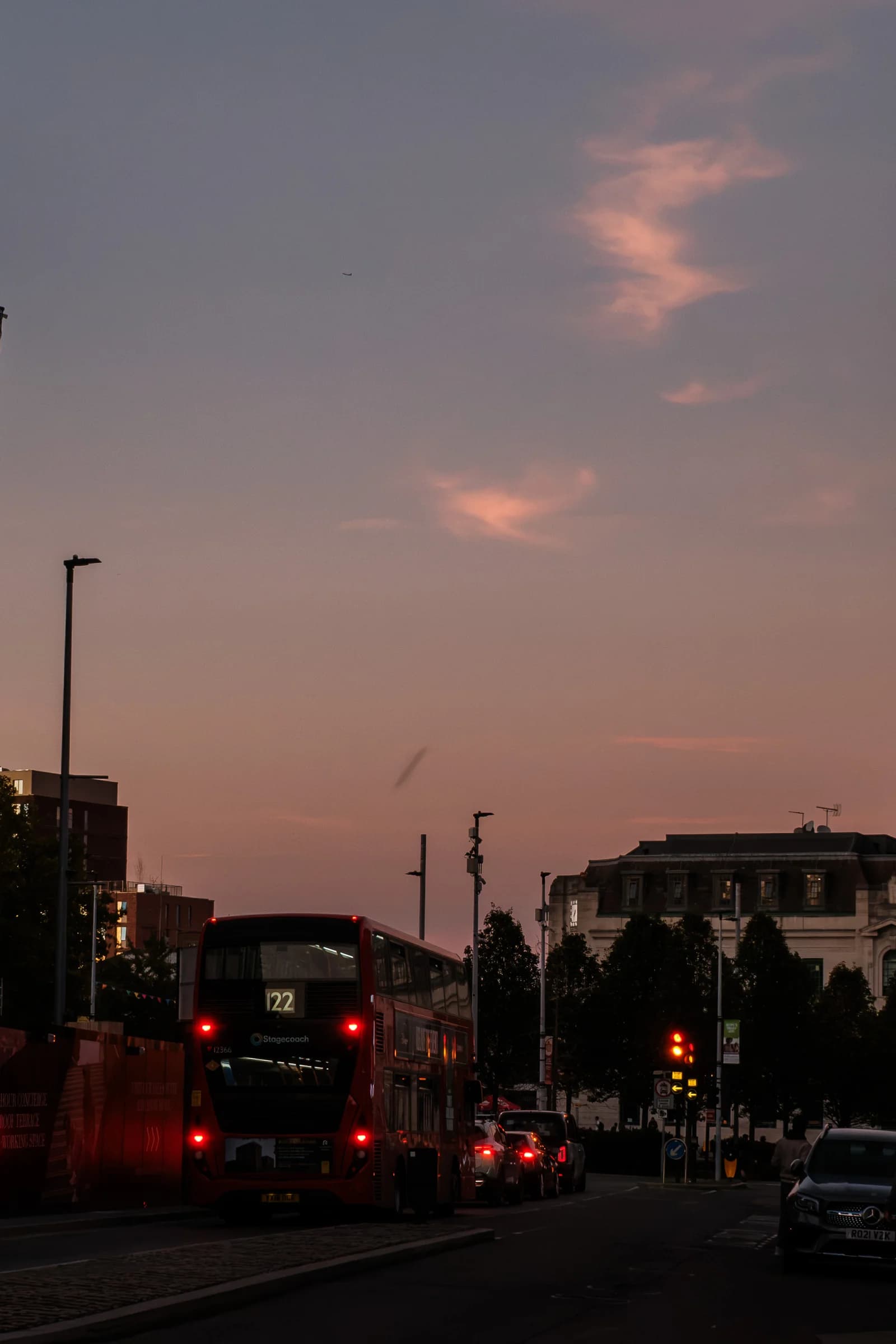 A red double-decker bus waits on a city street at twilight with its rear lights illuminated. Pink clouds are visible in the purple evening sky above buildings.