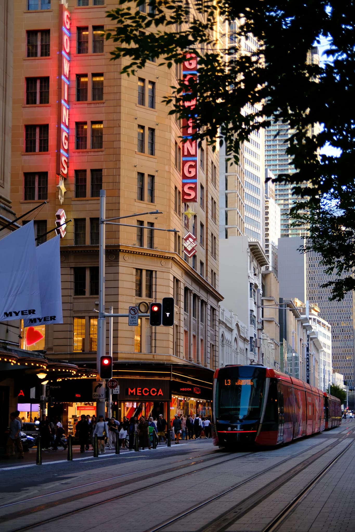 A red light rail tram travels down George Street in Sydney at dusk. Pedestrians walk past the historic Gowings building and brightly lit storefronts.