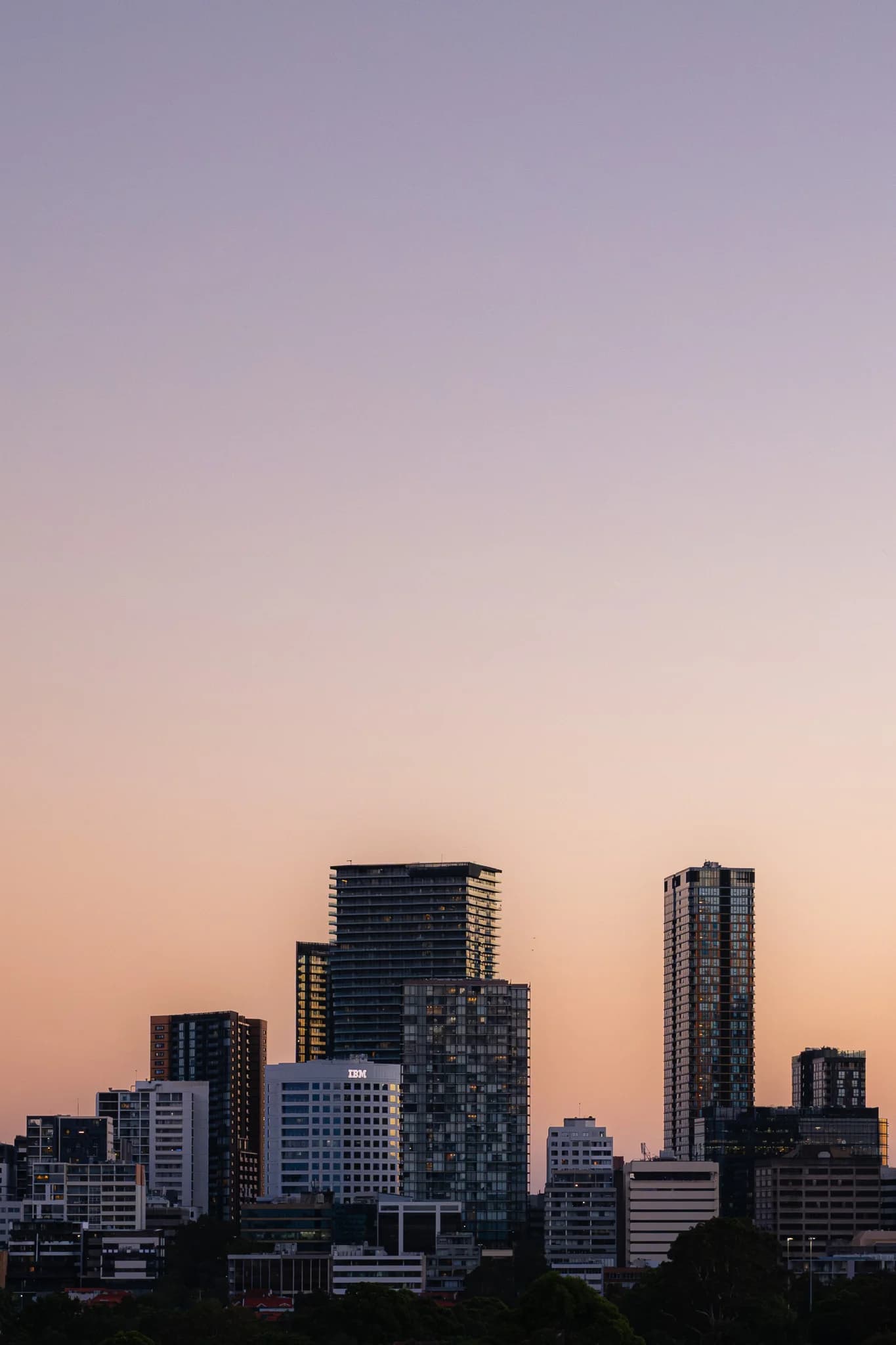 A modern city skyline stands against a smooth, pastel-coloured sky during dusk. Prominent high-rise buildings, including one with an IBM logo, are visible in the fading light.